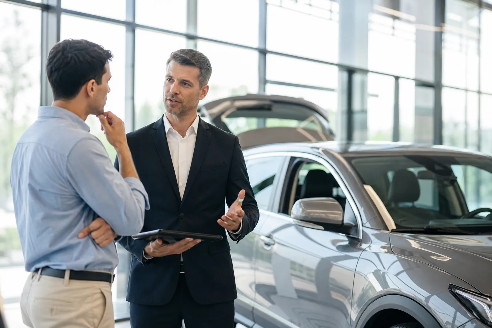 Un client discute avec un vendeur devant une voiture neuve dans un showroom lumineux.