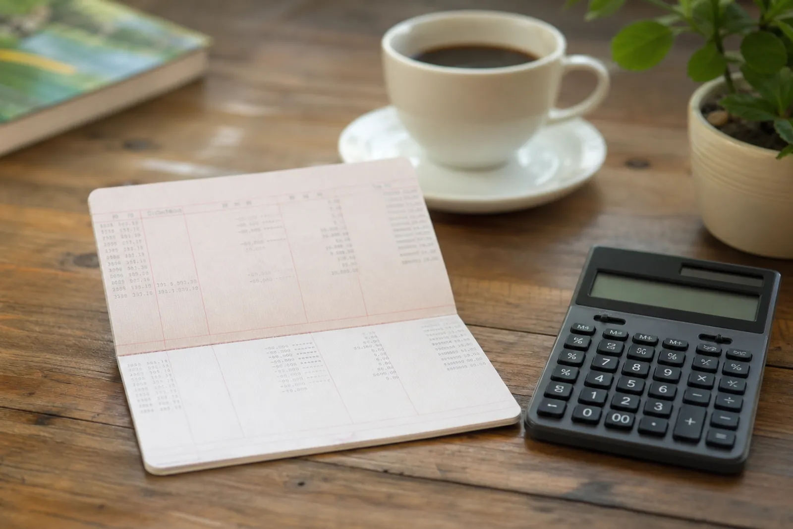Un carnet d'épargne et une calculatrice posés sur une table, avec une tasse de café en arrière-plan.